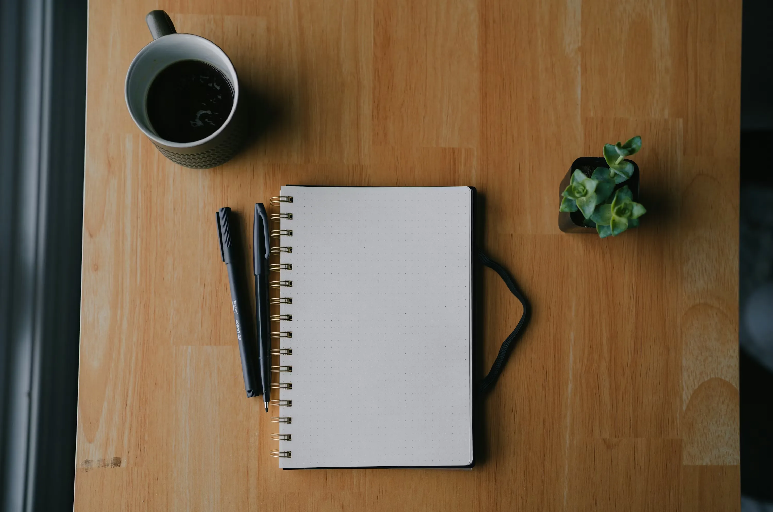 A notebook with a hand-drawn weekly meal schedule placed next to a cup of coffee on a wooden desk